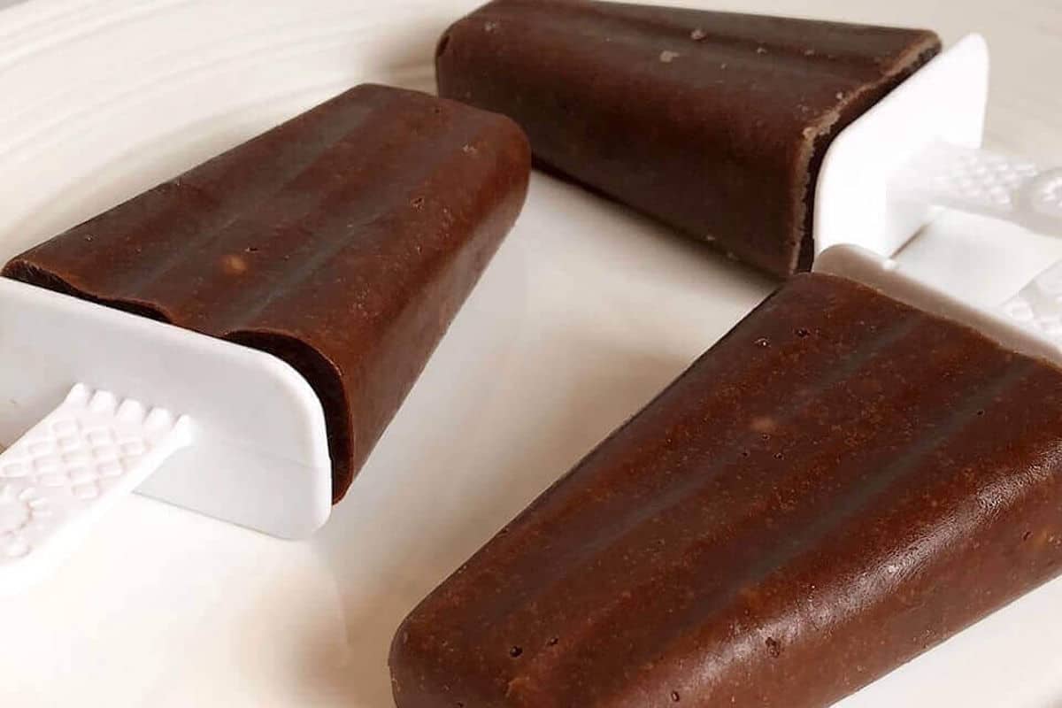Three homemade chocolate popsicles with white handles rest on a white plate.