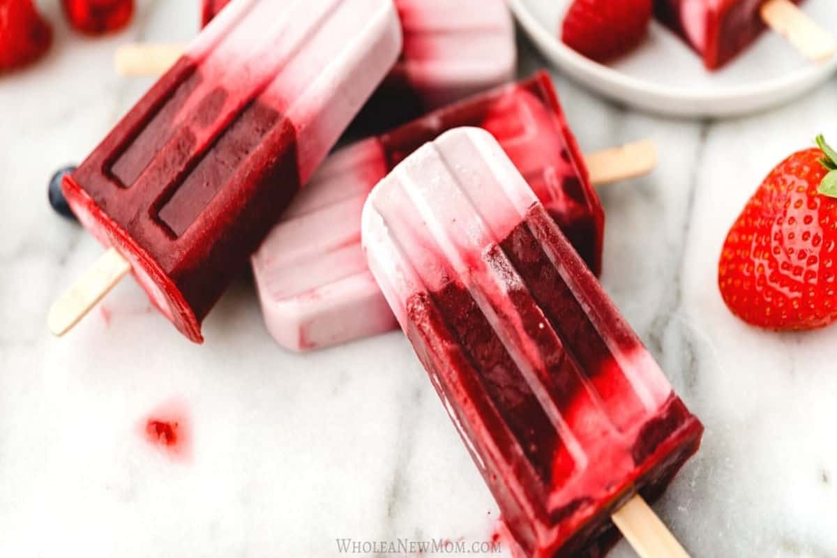 Mixed berry and coconut milk popsicles with a vibrant red and white gradient, arranged on a marble surface with fresh strawberries and raspberries.