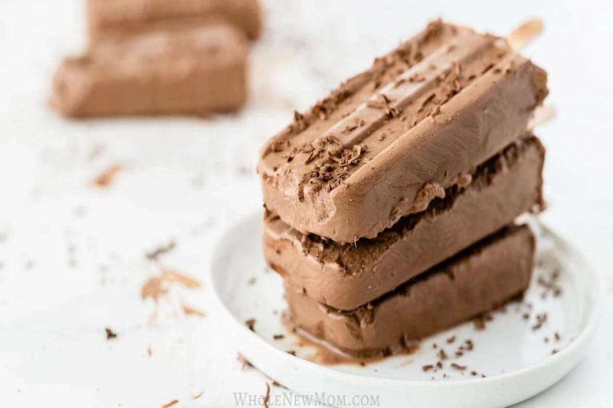 Stack of creamy chocolate fudgesicle popsicles sprinkled with chocolate shavings on a white plate, with more popsicles and shavings in the background.