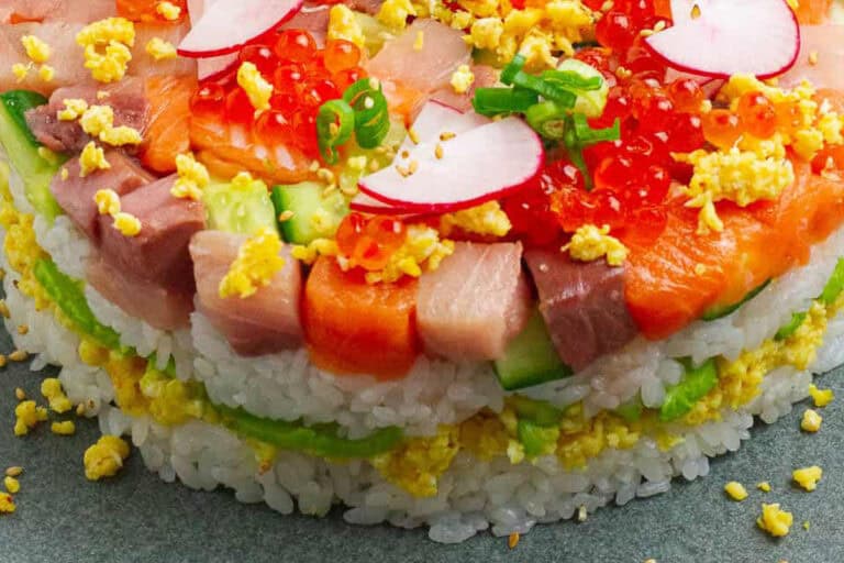A beautifully layered sushi cake with sushi rice, fresh salmon, avocado, and garnishes, served on a plate.