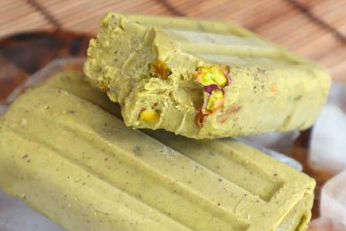 Green pistachio-flavored popsicles with a creamy texture, garnished with chopped pistachios, placed on ice in a wooden tray.
