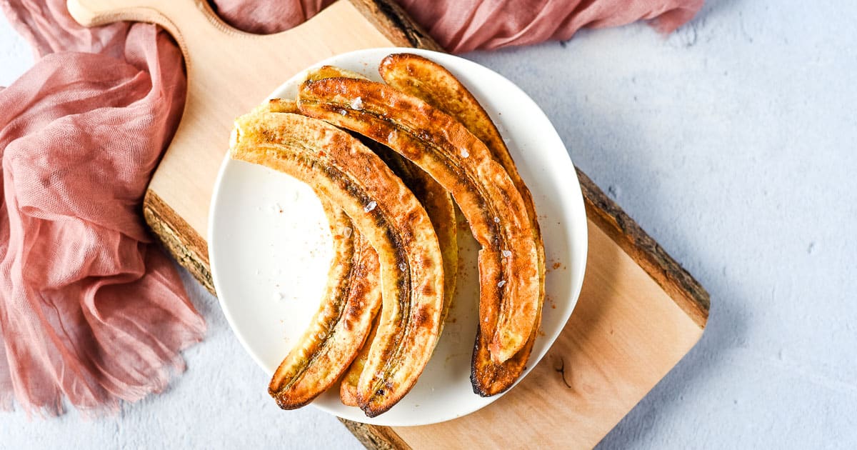 Air fryer bananas on a white plate over a rustic wooden cutting board with a pink linen.