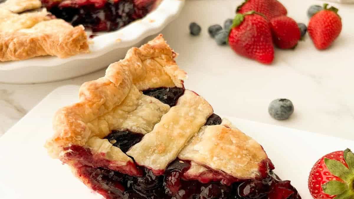 A slice of blueberry strawberry pie with a flaky golden lattice crust and glossy berry filling, served alongside fresh strawberries and blueberries.
