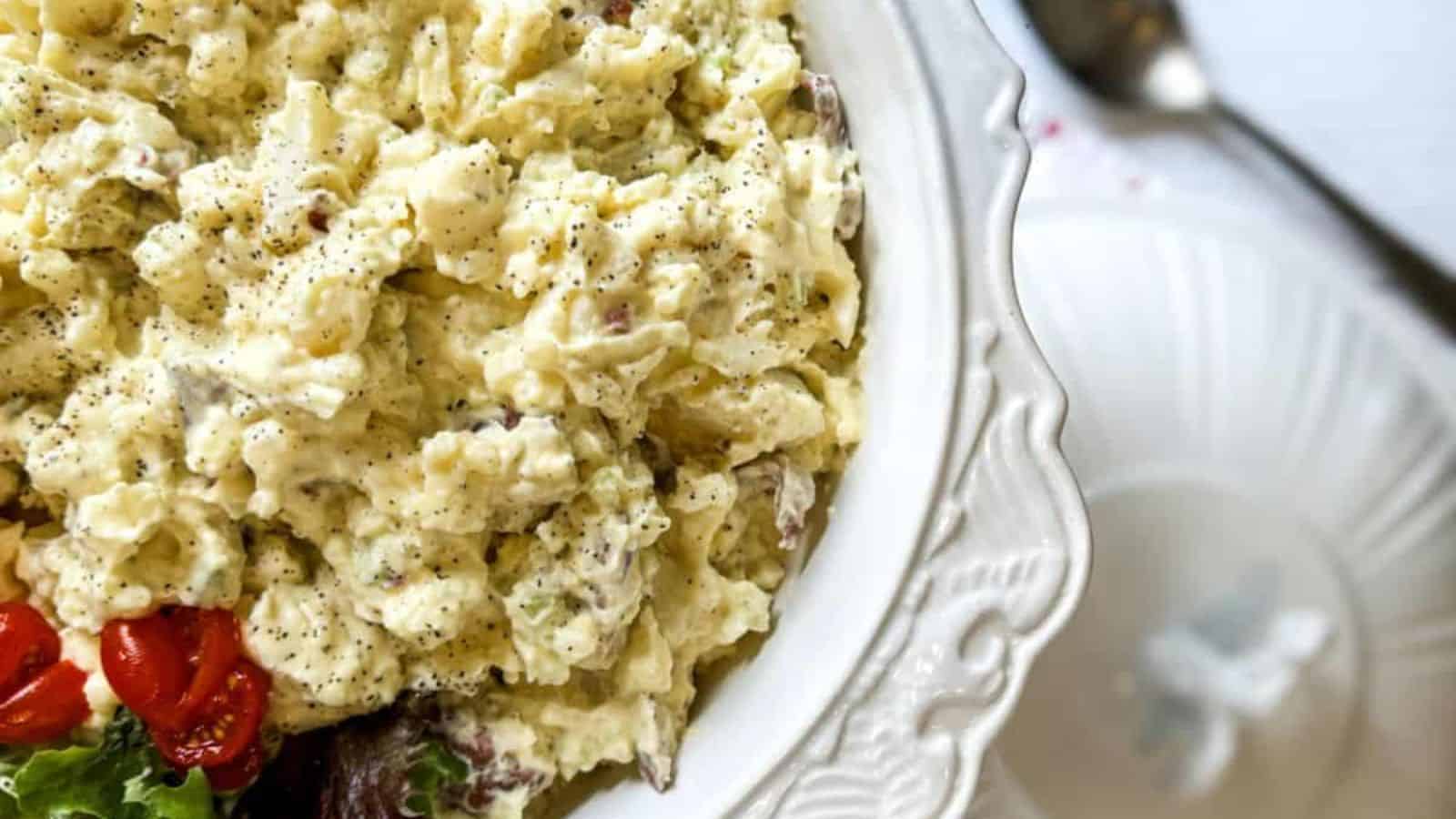 A bowl of creamy potato salad with specks of pepper, garnished with cherry tomatoes and lettuce on a decorative white plate.