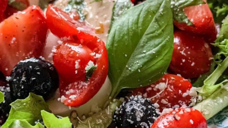 Close-up of a fresh salad featuring strawberries, cherry tomatoes, blueberries, and basil leaves on a bed of mixed greens. The salad is sprinkled with grated cheese, adding a textured contrast to the vibrant colors of the fruits and vegetables.
