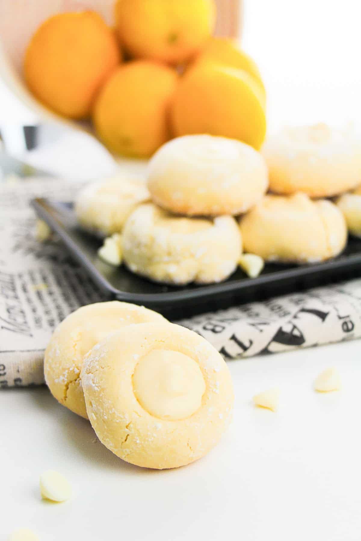 A plate of lemon cookies displayed on a newspaper, with lemons and white chocolate chips nearby.