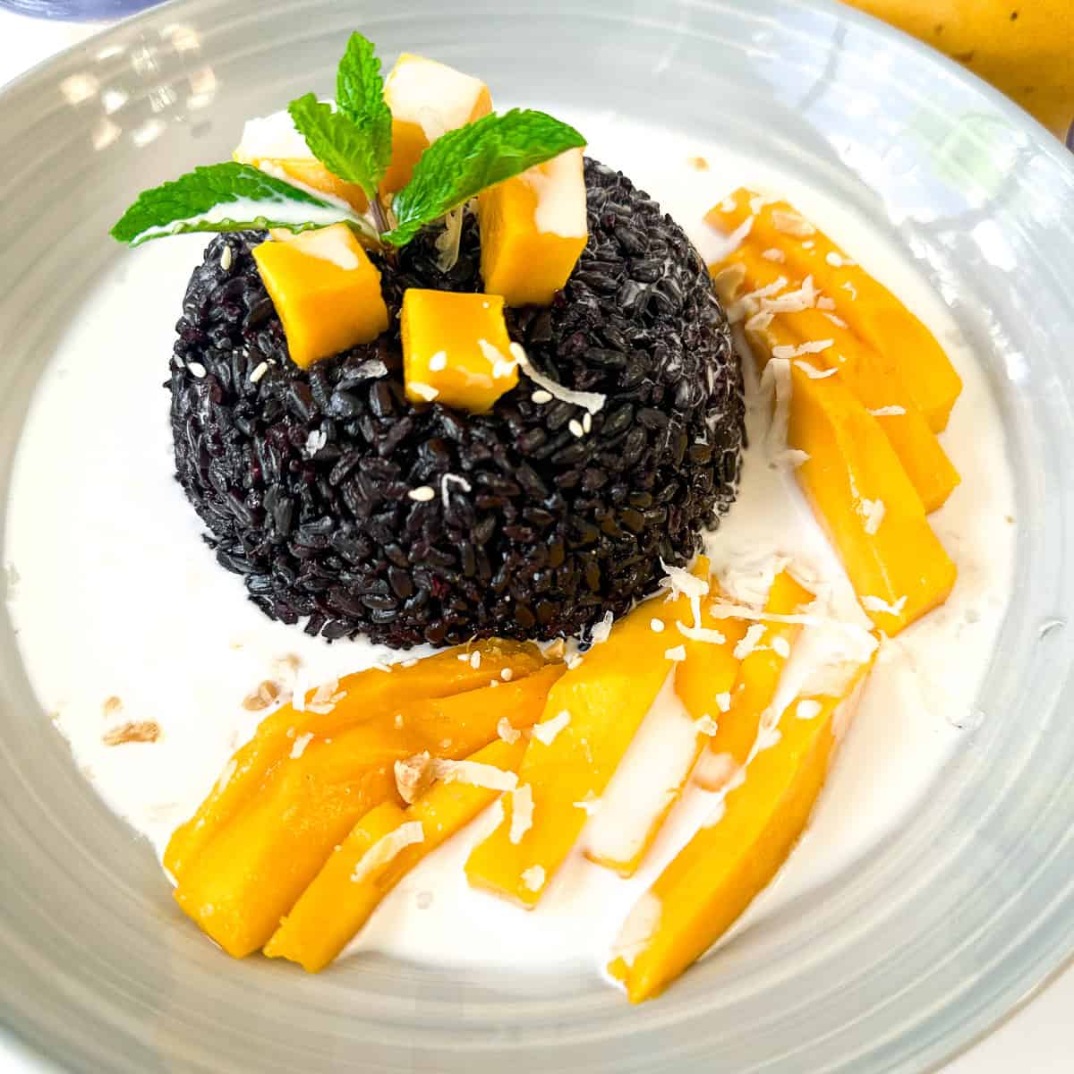 A bowl of black sticky rice pudding garnished with mango slices and sprinkled with coconut flakes.