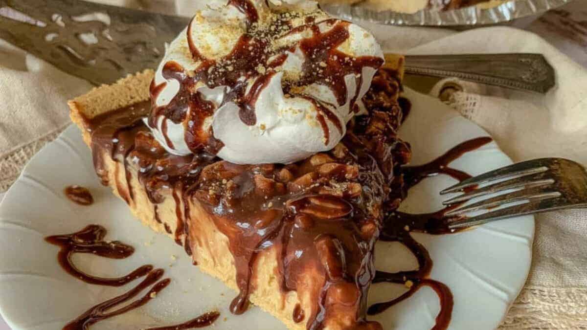 A slice of pie with chocolate drizzle and whipped cream is presented on a white plate with a fork.