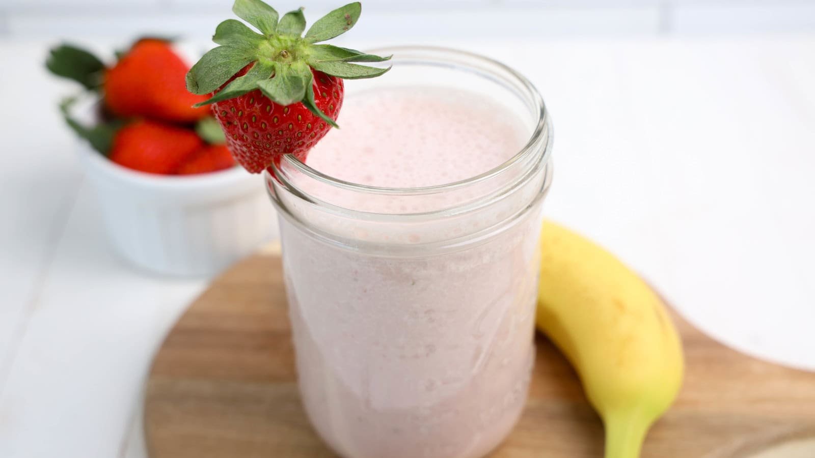 A strawberry banana smoothie in a glass, with a strawberry on top for decoration on a cutting board.