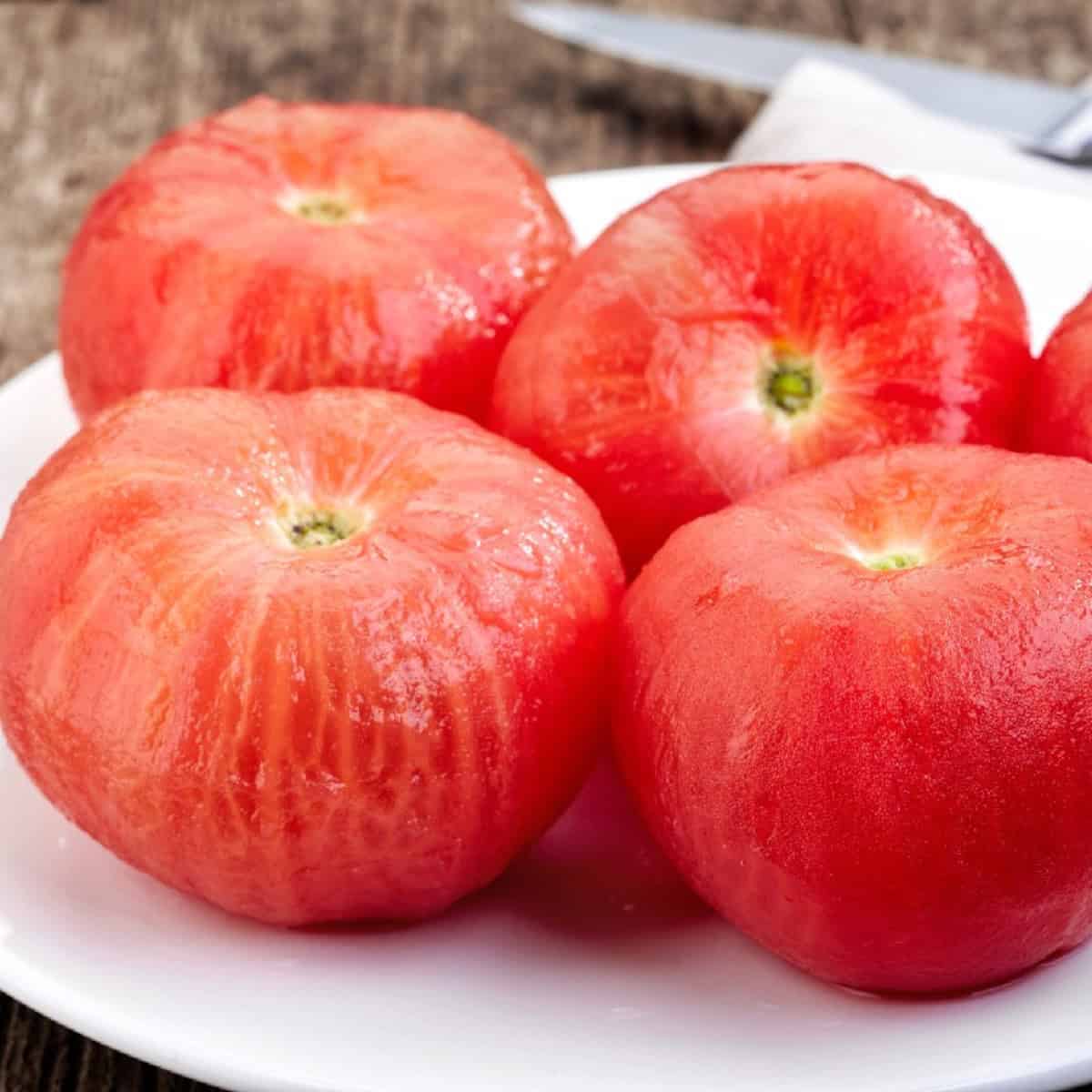 A plate holding four ripe, red tomatoes with the skin peeled off.