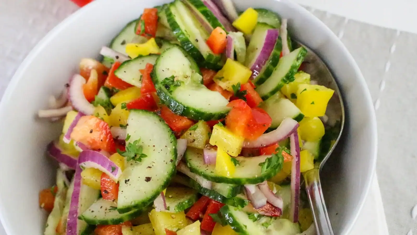 A bowl of cucumber salad with red onion, bell peppers, tomatoes, and herbs, served with a spoon.
