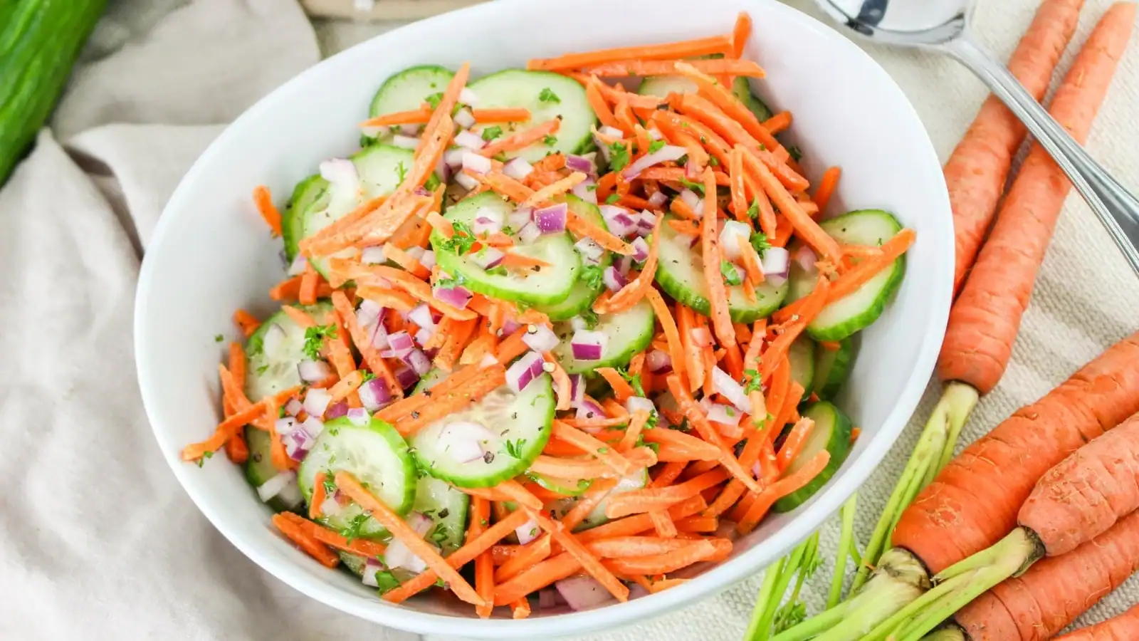 A white bowl contains a salad made of sliced cucumbers, shredded carrots, and diced red onions. Fresh carrots are placed beside the bowl.