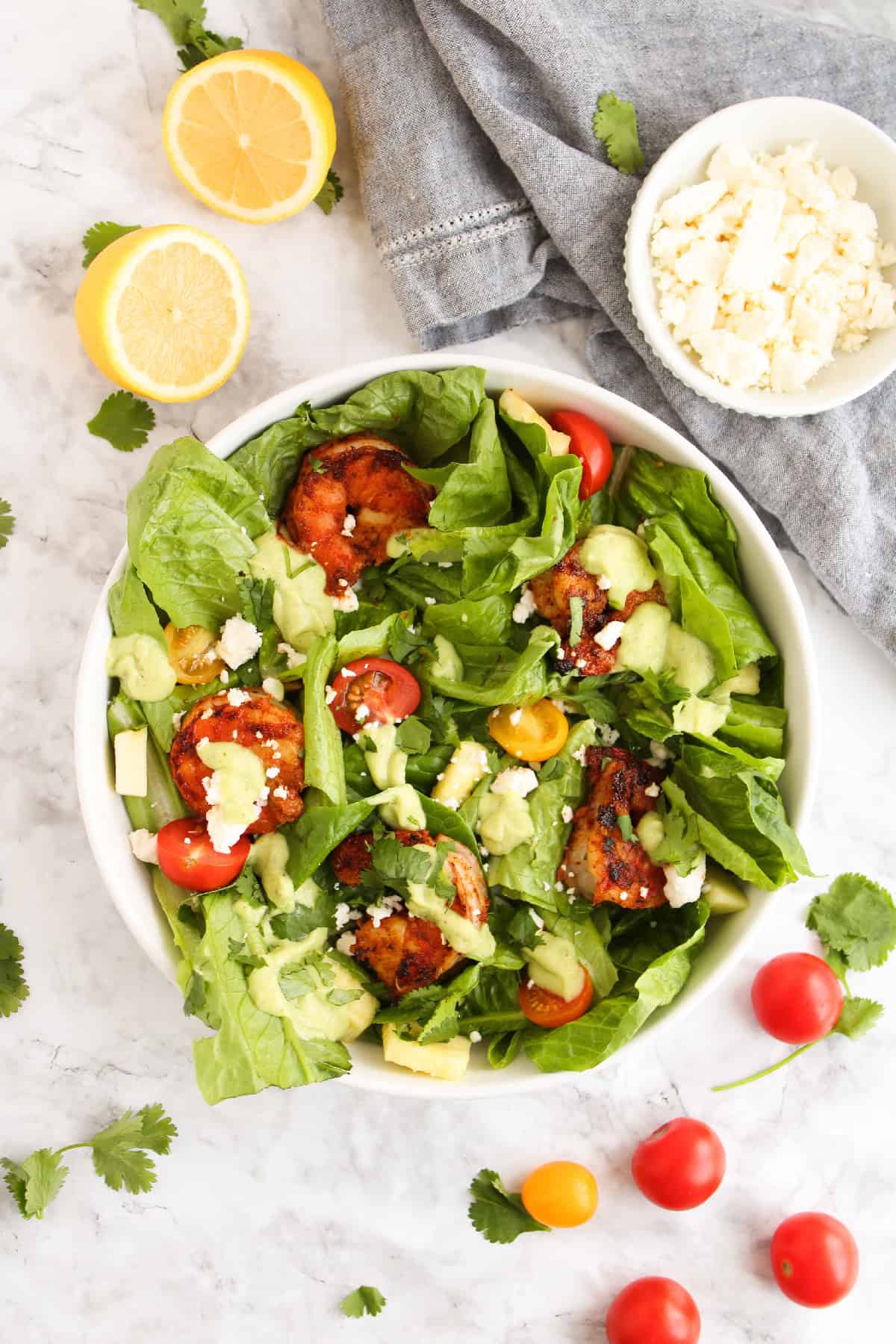 Close-up of a blackened shrimp salad with avocado dressing, cherry tomatoes, fresh romaine, feta cheese, and cilantro in a white bowl.