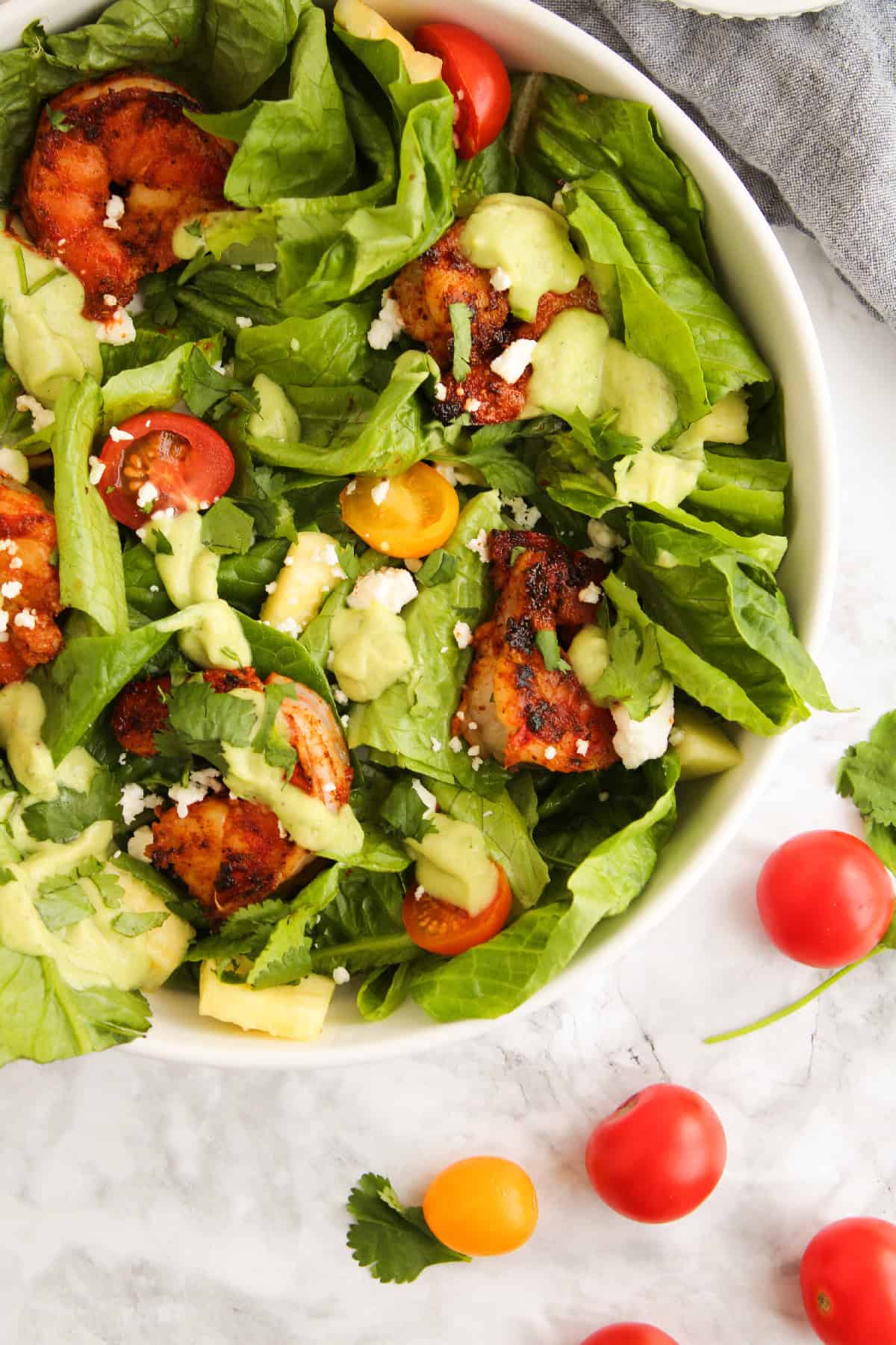 Close-up of a blackened shrimp salad with avocado dressing, cherry tomatoes, fresh romaine, feta cheese, and fresh cilantro in a white bowl.