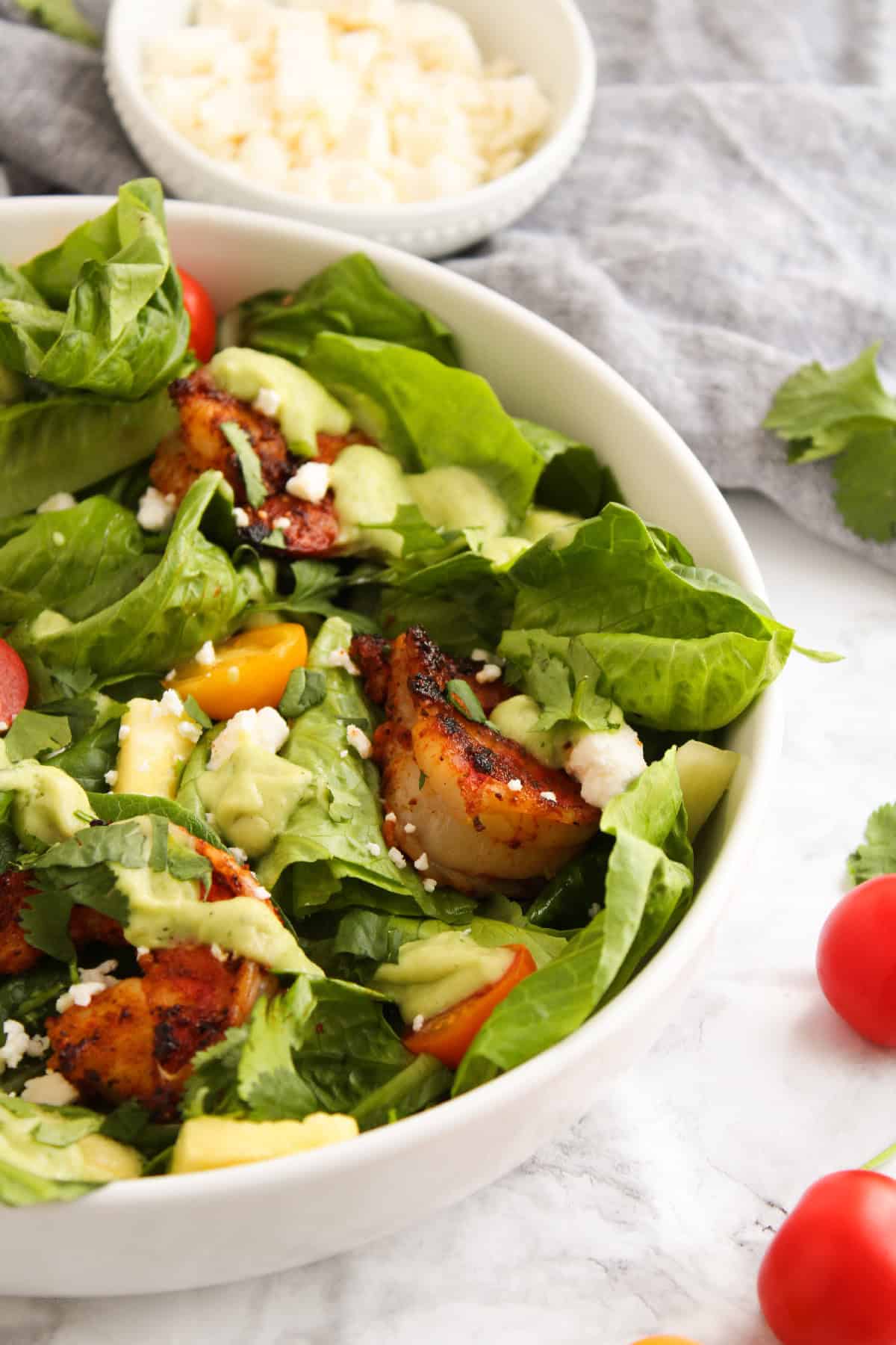 Close-up of a blackened shrimp salad with avocado dressing, red cherry tomatoes, fresh romaine, feta cheese, and cilantro in a white bowl.