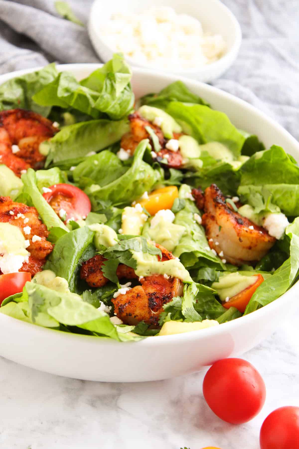 Close-up of a blackened shrimp salad with avocado dressing, cherry tomatoes, romaine, feta cheese, and cilantro in a white bowl.