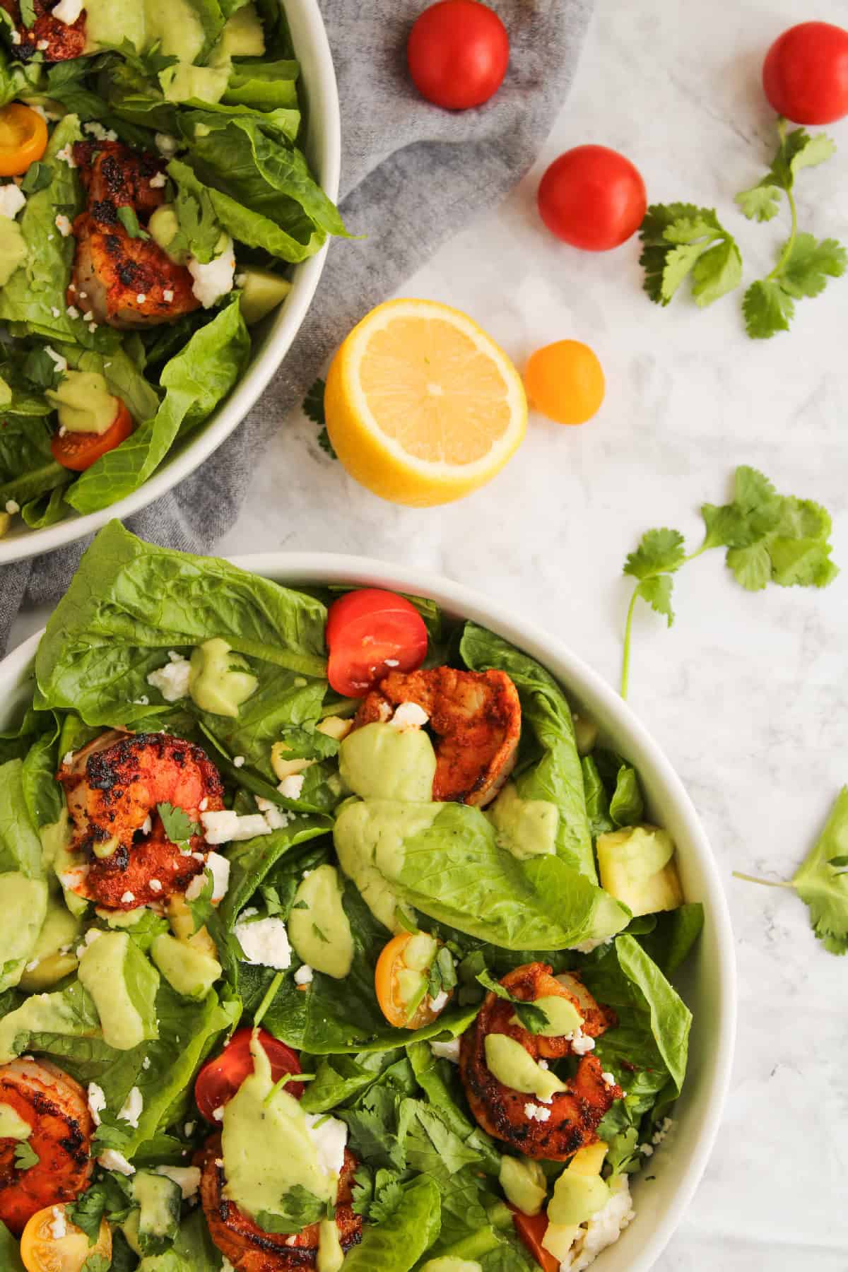 Close-up of two blackened shrimp salad with avocado dressing, cherry tomatoes, fresh romaine, feta cheese, and cilantro in a white bowl.
