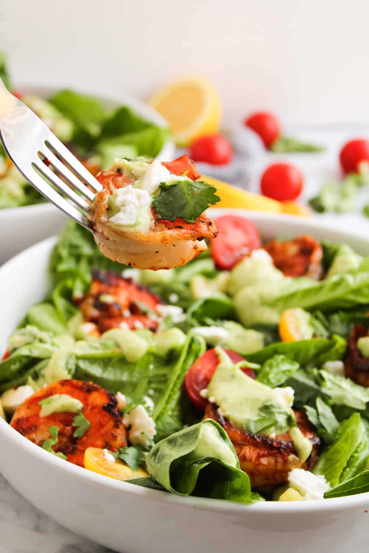 Fork holding a bite of blackened shrimp with avocado dressing and feta cheese over a bowl of fresh shrimp salad with tomatoes and romaine.