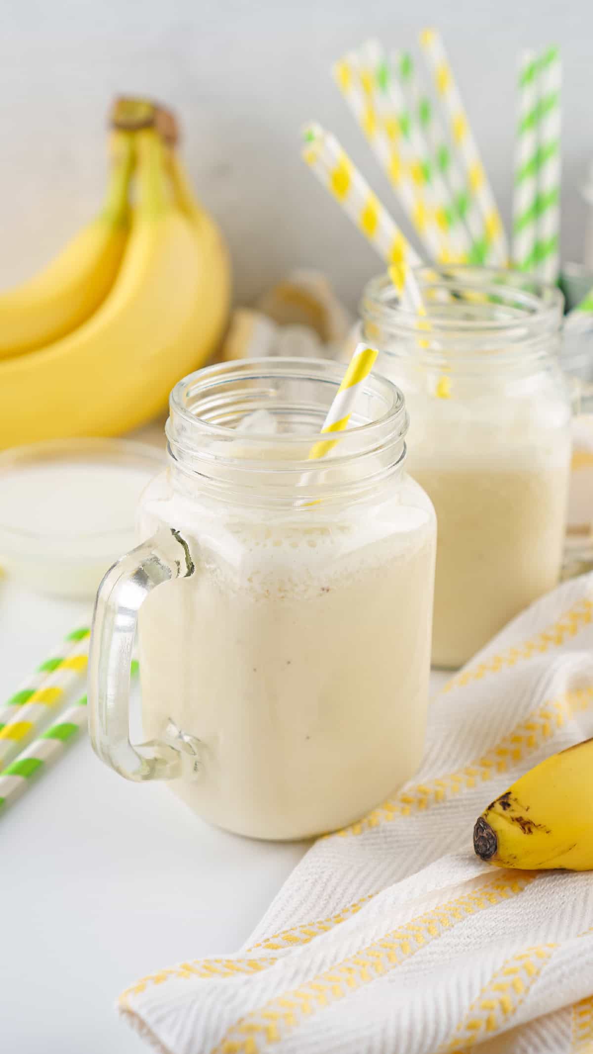 Banana milk in mason jars served on a kitchen counter with fresh bananas, straws, and a striped towel.