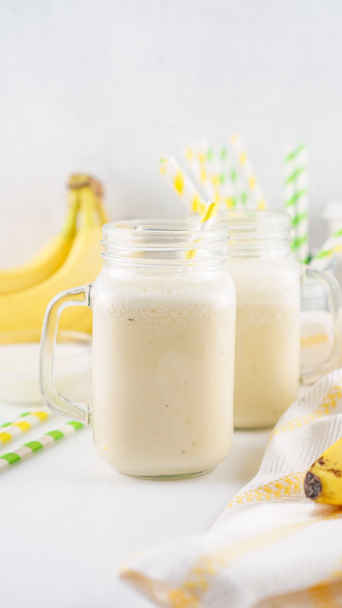 Two chilled jars of banana milk with straws, bananas, and colorful straws in the background.