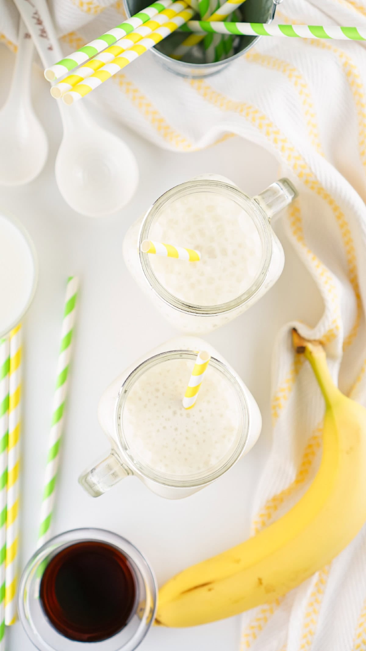 Top view of two jars of banana milk with yellow paper straws on a white surface with green and yellow accents.