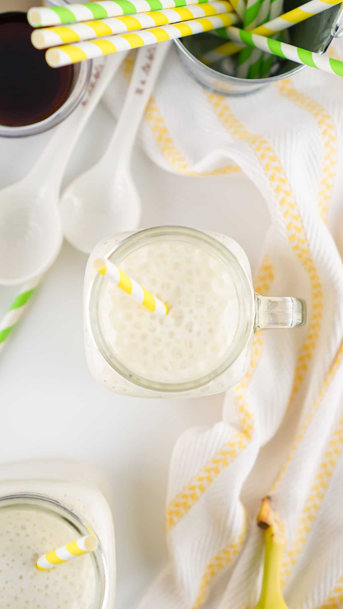 Top view of banana milk in mason jar with yellow straw, surrounded by striped paper straws and a kitchen towel.