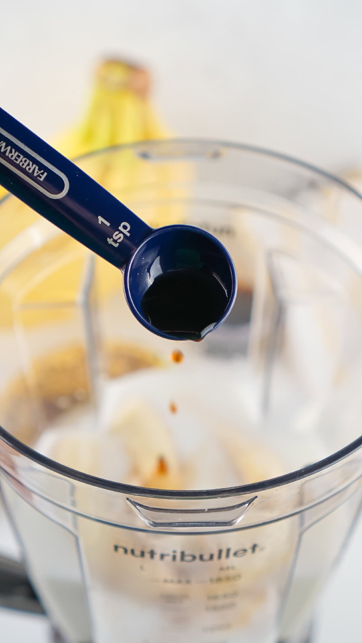 Vanilla extract in a blue measuring spoon added to a banana milkshake blend.