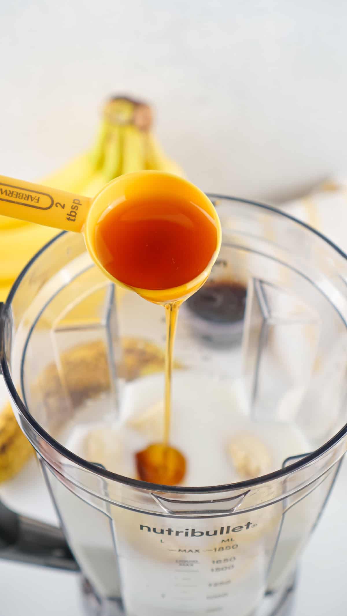 Maple syrup measured in a yellow spoon being poured into banana milk mixture.