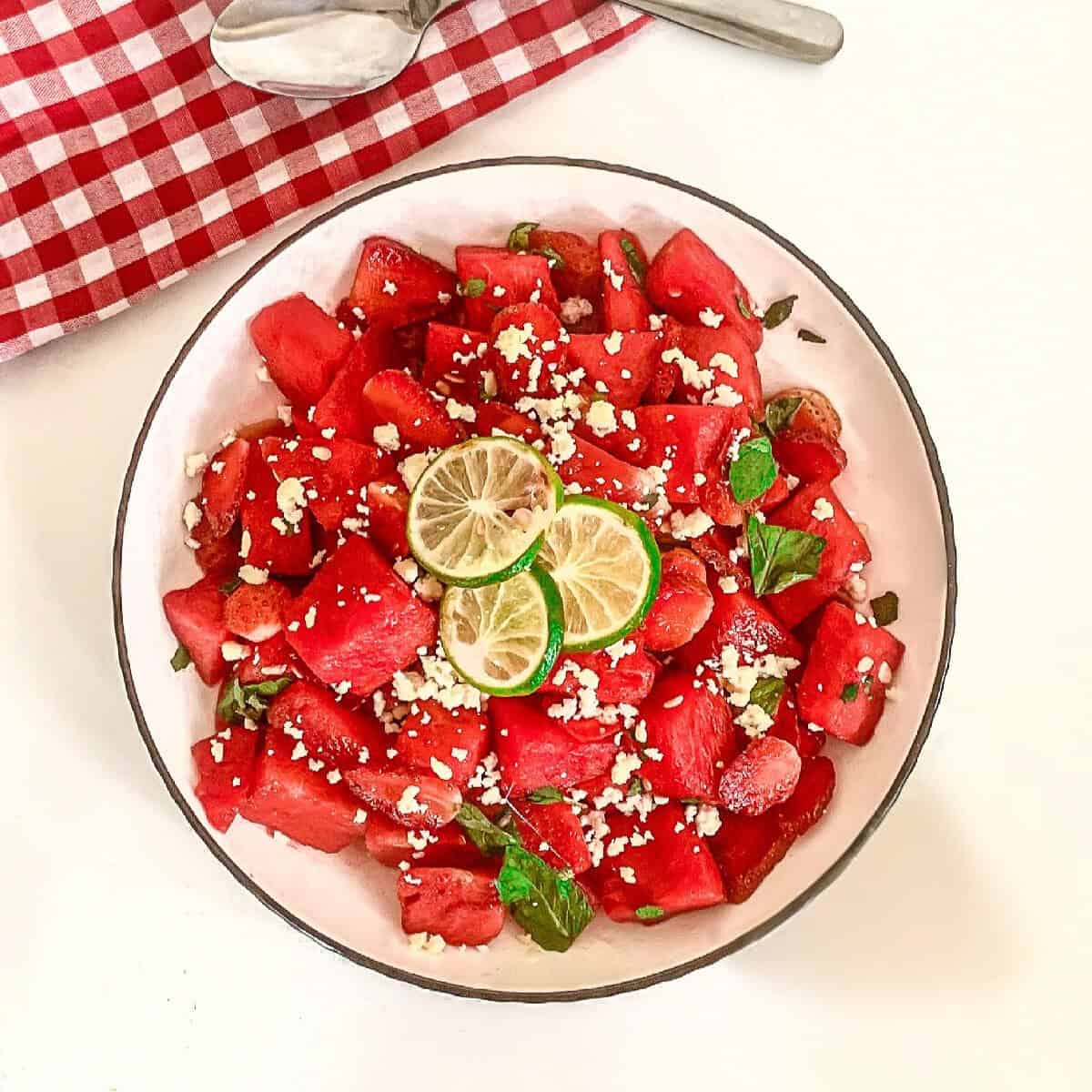 Bowl of fresh watermelon strawberry salad topped with feta cheese, lime slices, and mint leaves on a white background with red gingham napkin.