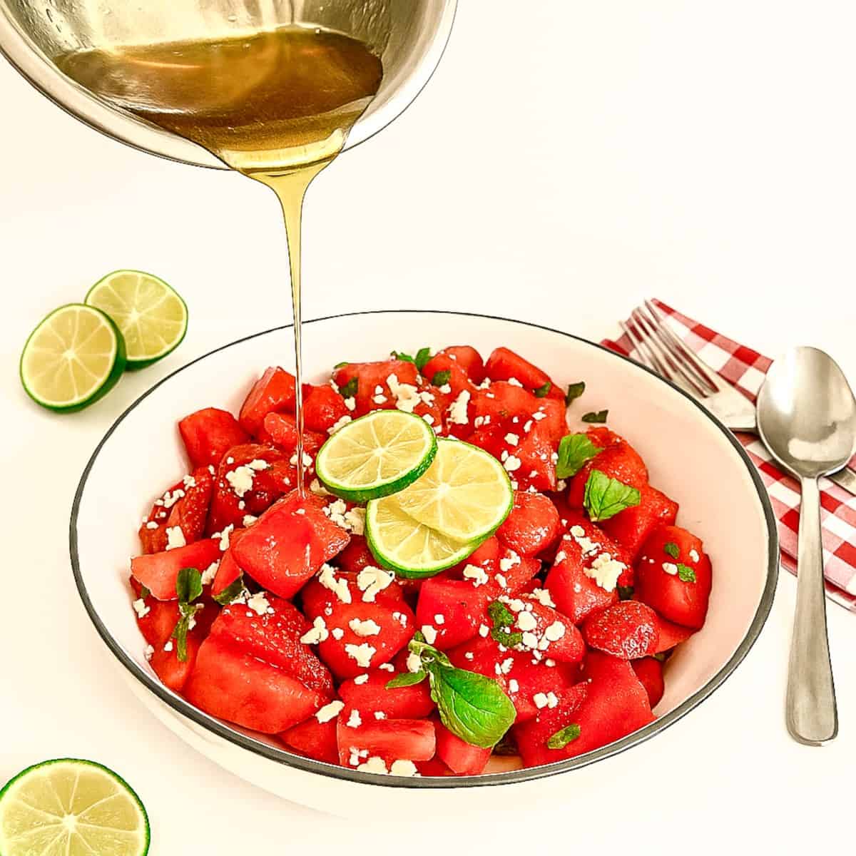 Honey-lime dressing being poured onto watermelon strawberry salad with feta and mint in a white bowl, styled with lime slices and red gingham napkin.