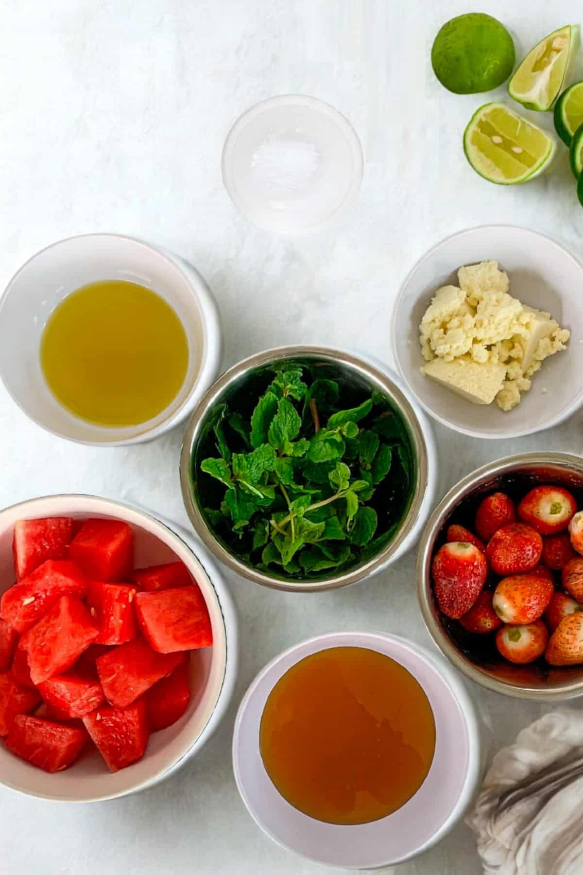 Flat lay of ingredients for watermelon strawberry salad including watermelon cubes, strawberries, mint, honey, olive oil, feta cheese, lime, and salt.