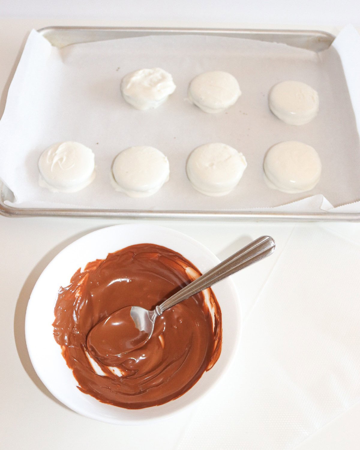 Melted chocolate in a bowl with a spoon, ready for decorating the white chocolate-covered Oreo cookies on the tray.