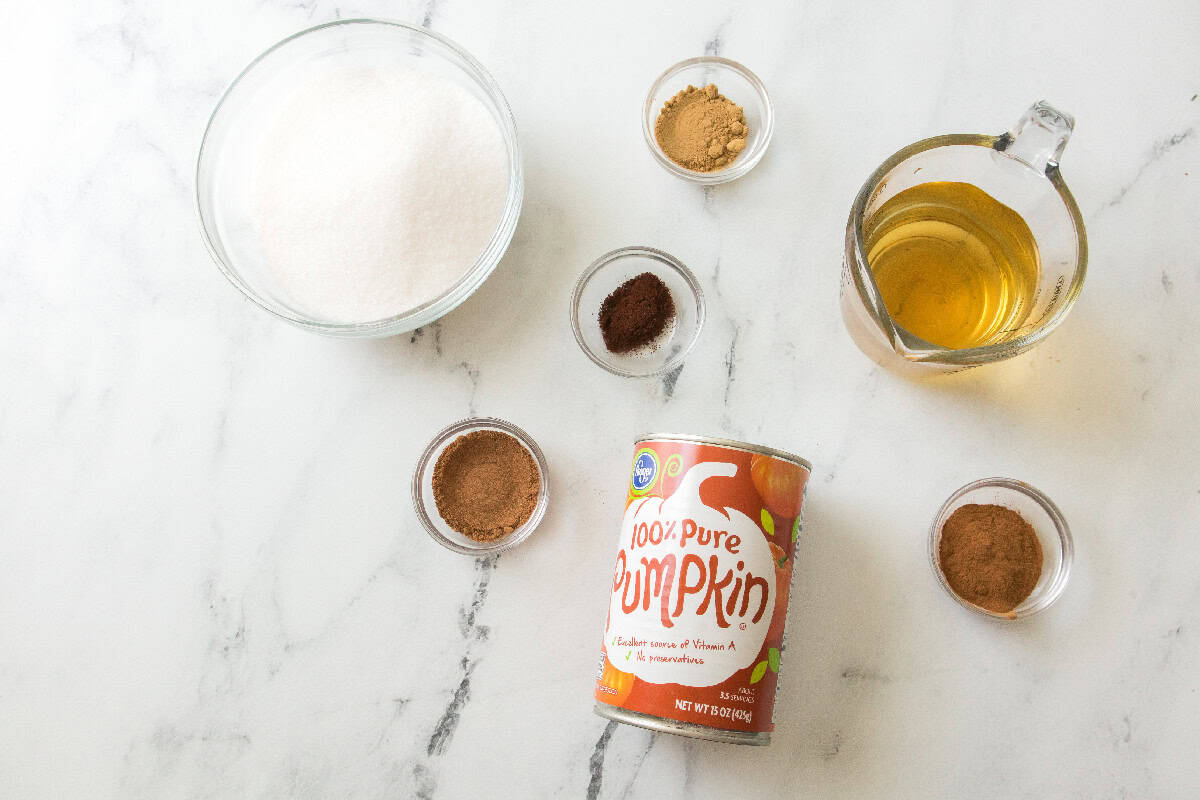 Overhead view of pumpkin butter ingredients on a white marble surface, including a can of pure pumpkin, a bowl of white sugar, a measuring cup of apple juice, and small glass bowls filled with ground cinnamon, ginger, cloves, and nutmeg.