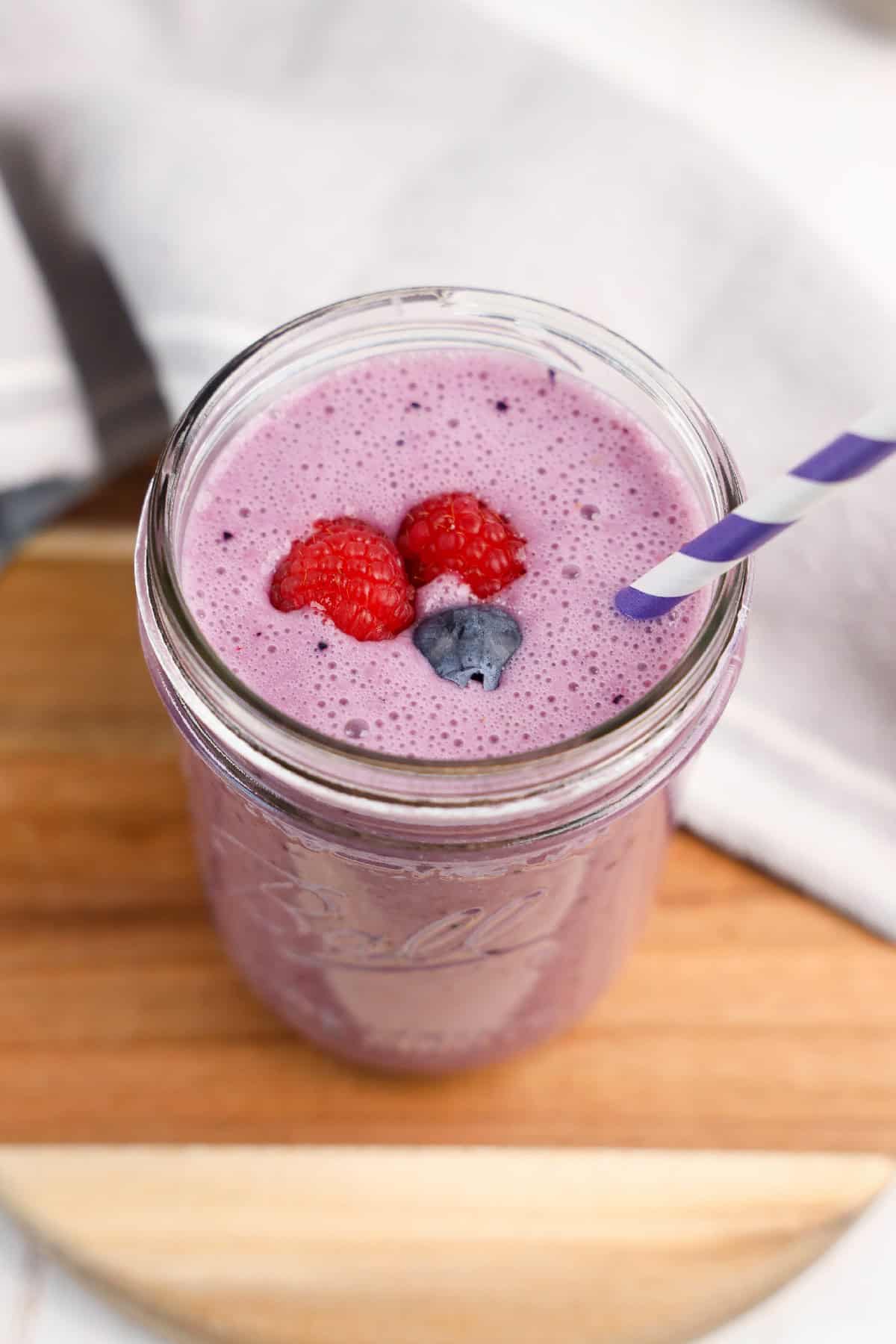 Overhead view of a purple vegan triple berry smoothie in a mason jar, topped with fresh raspberries and a blueberry, with a purple-striped straw on a wooden board.