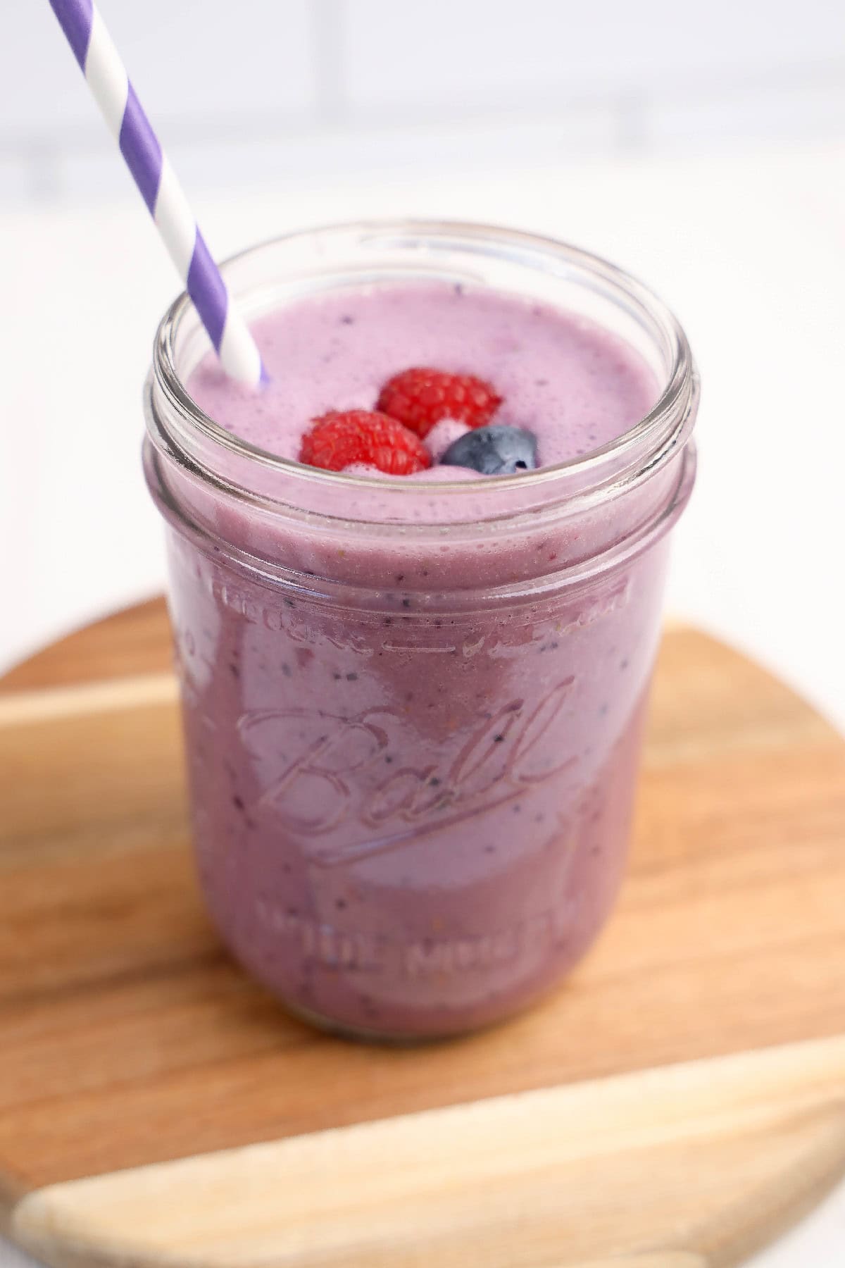 Overhead view of a vibrant purple vegan triple berry smoothie in a mason jar, topped with fresh raspberries and a blueberry, with a purple-striped straw.