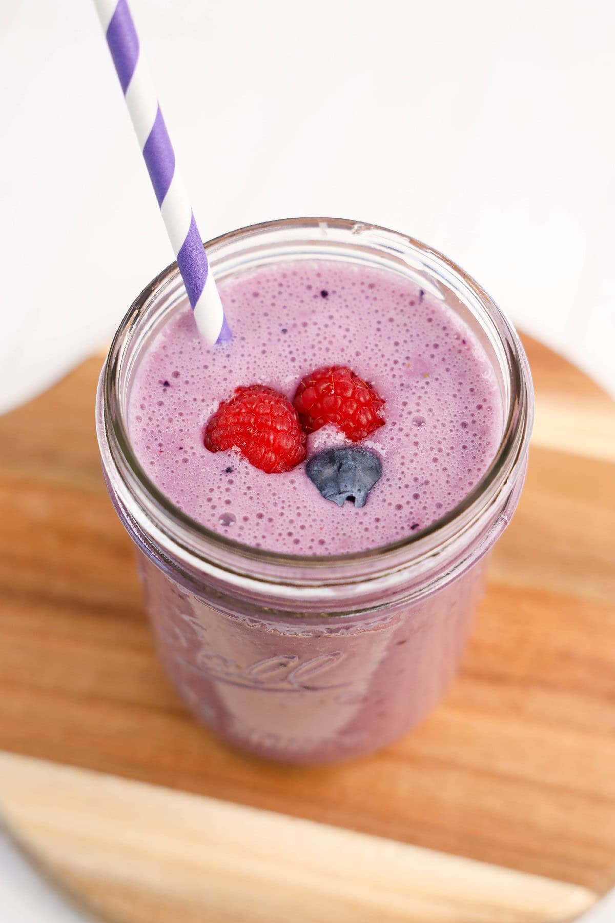 Overhead view of a vibrant purple vegan triple berry smoothie in a mason jar, topped with fresh raspberries and a blueberry, with a straw on a wooden board.