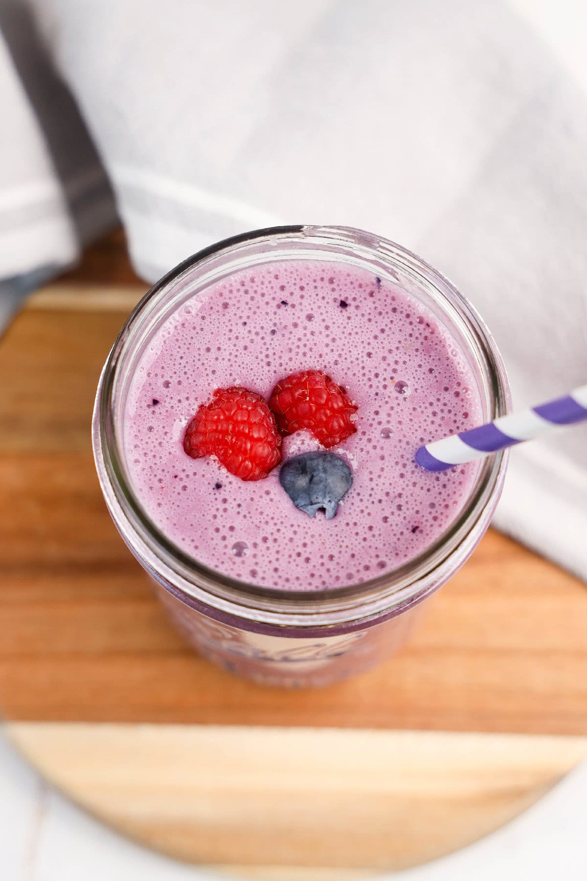 Overhead view of a vibrant purple vegan triple berry smoothie in a mason jar, topped with fresh raspberries and a blueberry, with a purple-striped straw on a wooden board.
