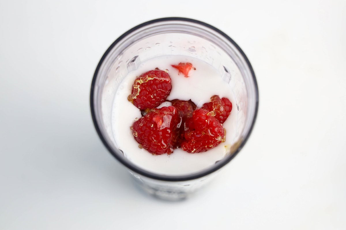 Fresh raspberries and a drizzle of honey floating in coconut milk inside a blender cup.