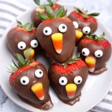 Chocolate-covered strawberries decorated with candy eyes and candy corn to look like cute Thanksgiving turkeys, arranged on a white plate.