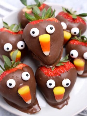 Chocolate-covered strawberries decorated with candy eyes and candy corn to look like cute Thanksgiving turkeys, arranged on a white plate.