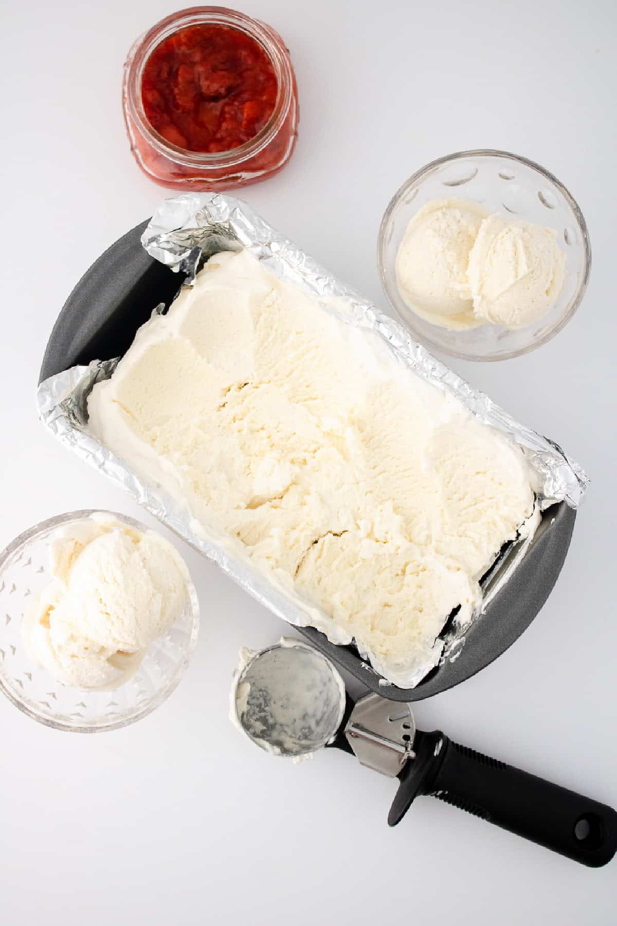 Pan of vanilla bean no-churn ice cream with bowls of scoops and jar of strawberry sauce.