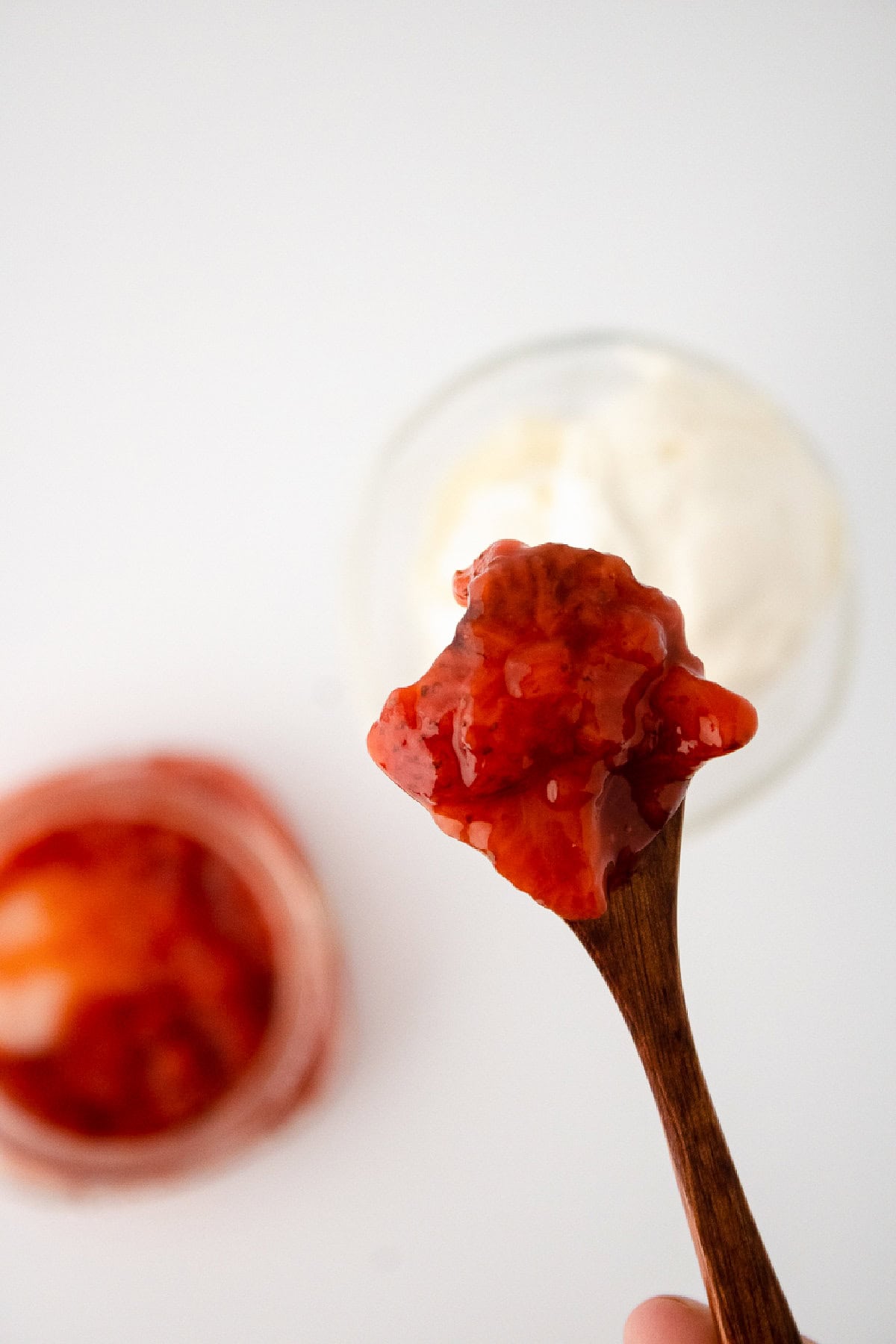 Wooden spoon holding chunky strawberry sauce over vanilla ice cream bowl.