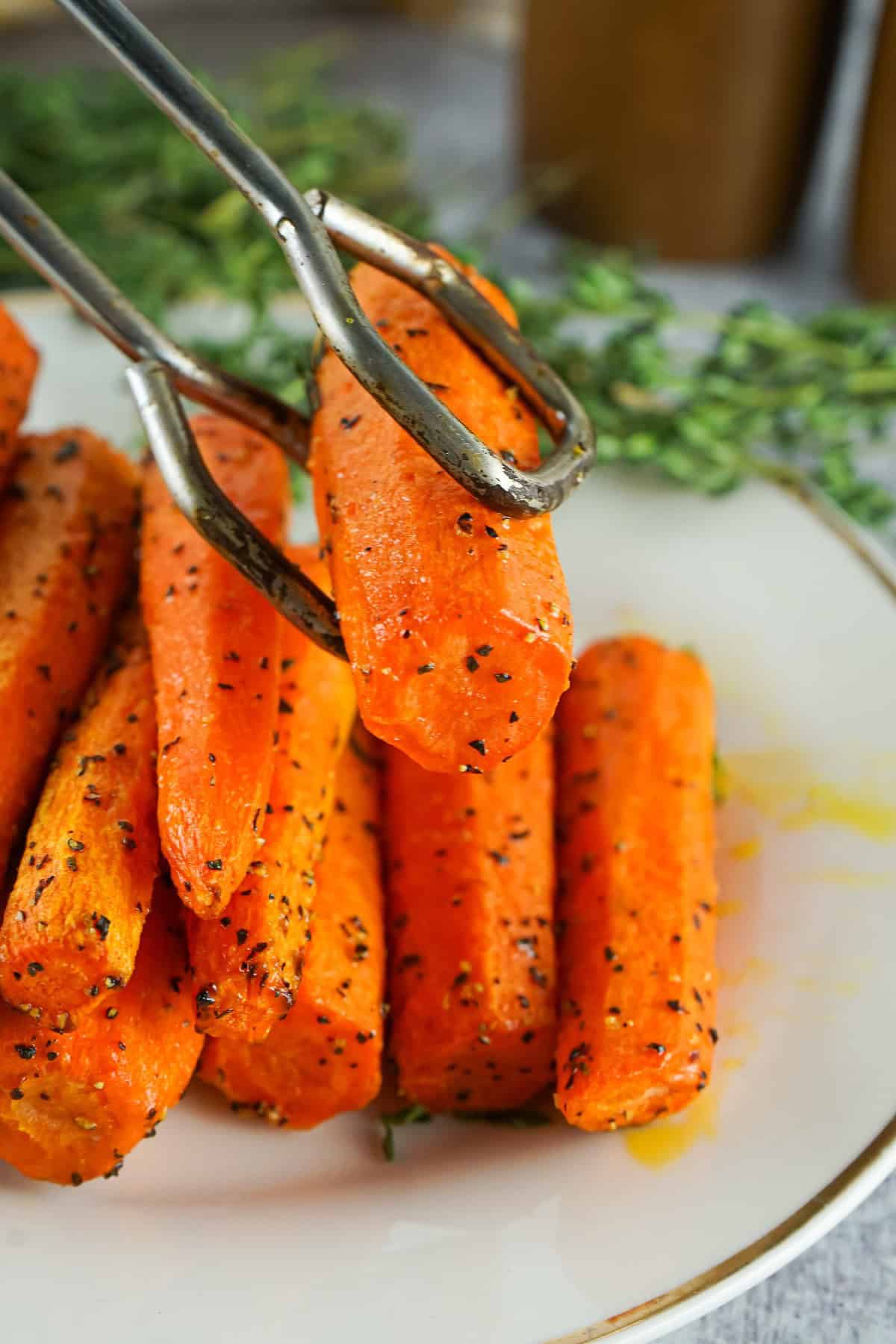 Metal tongs holding a roasted air fryer carrot over a plate of herb-seasoned carrots, ready to serve.