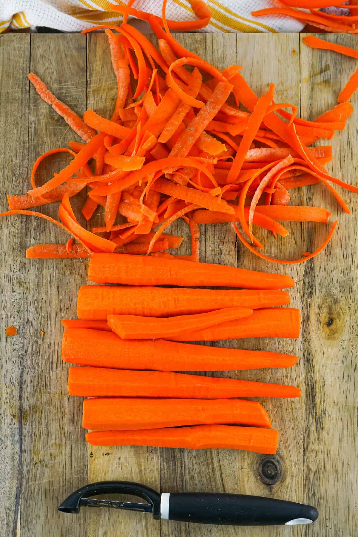 Peeled and sliced carrots on a wooden cutting board with vegetable peeler for air fryer cooking.
