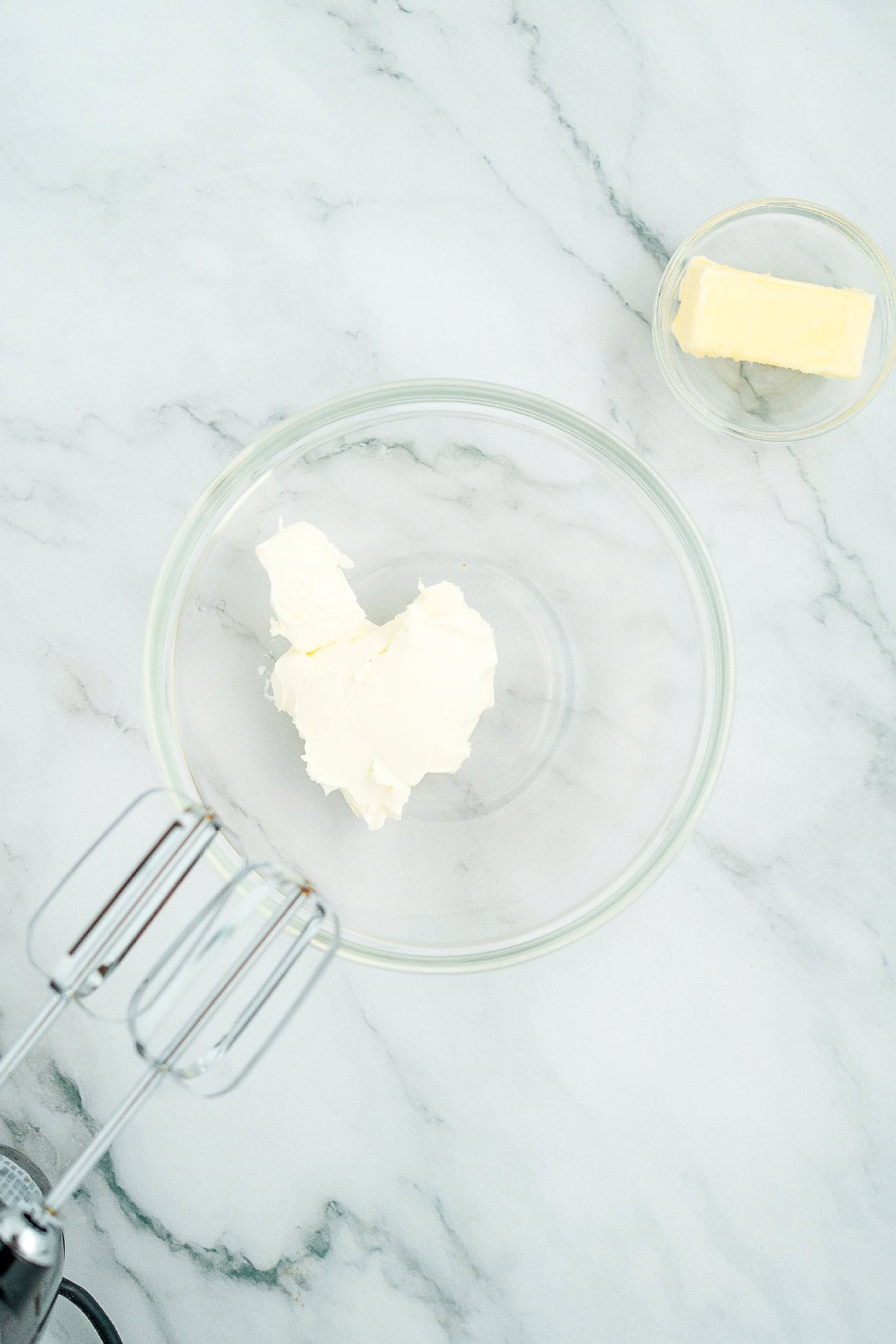 Bowl with cream cheese and butter on a marble countertop next to a hand mixer.