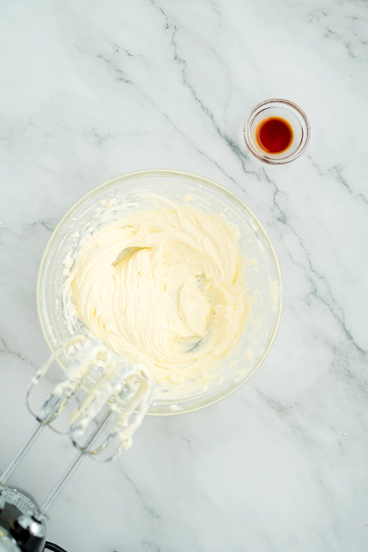 Creamy whipped mixture of cream cheese and butter in a glass bowl with vanilla extract on the side.