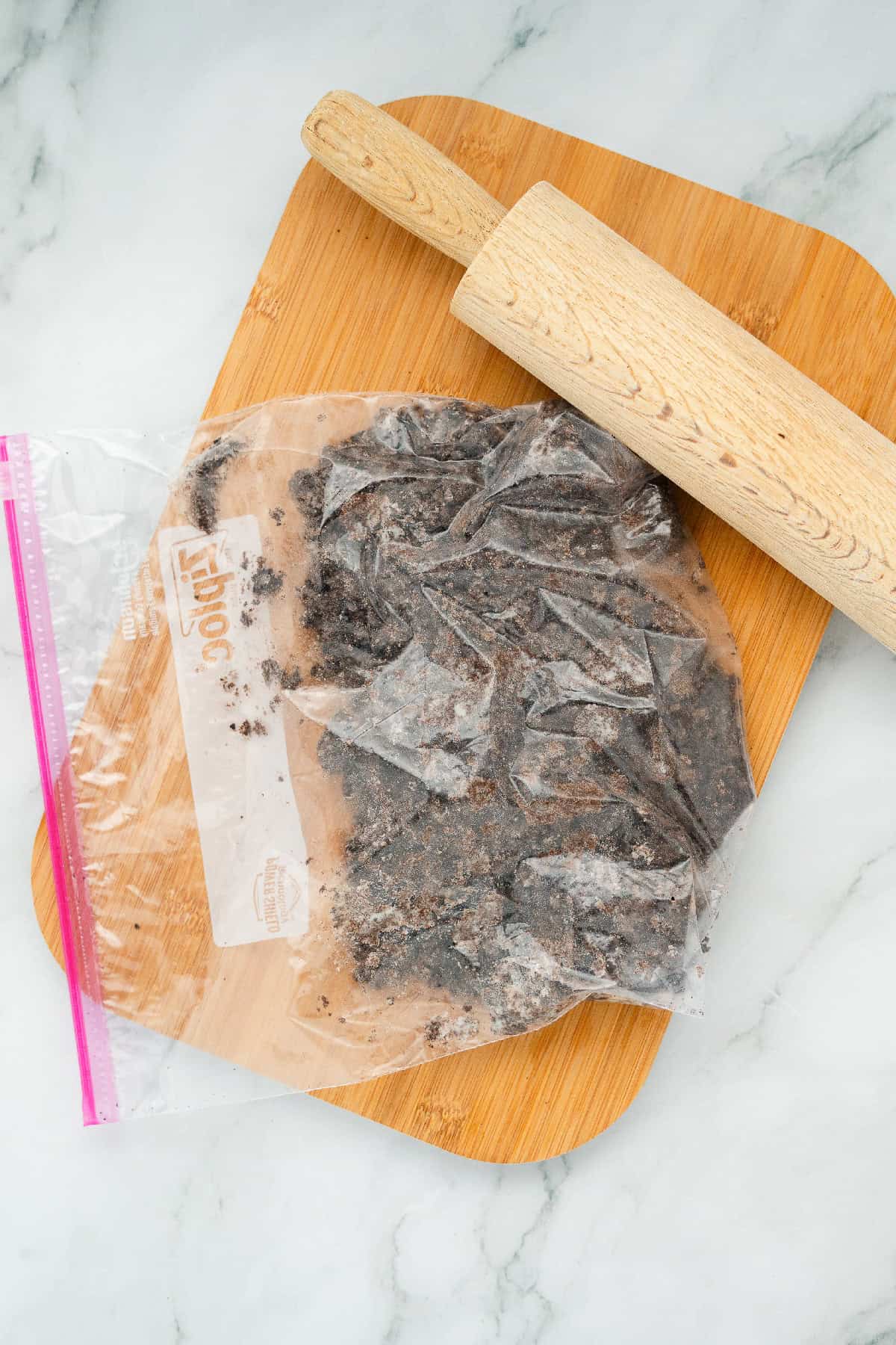 Crushed Oreo cookies inside a plastic bag with a rolling pin on a wooden cutting board for dirt cake cups.