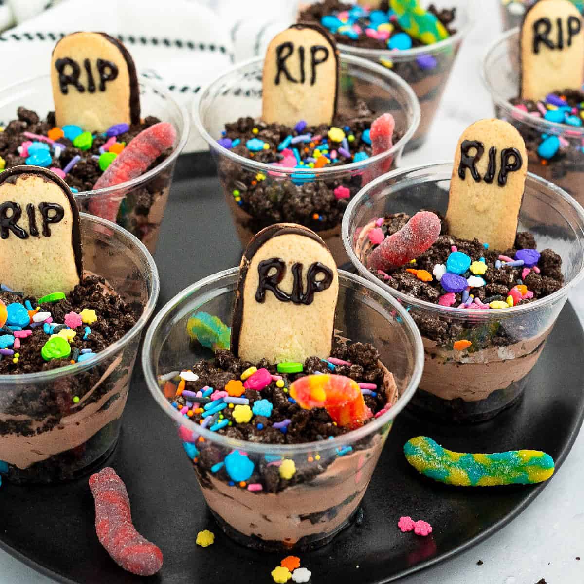 Spooky graveyard dirt pudding cups decorated with cookie tombstones, gummy worms, and bright Halloween sprinkles.