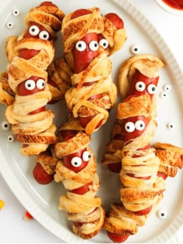 Hot dogs wrapped in crescent roll dough to look like mummies, baked golden brown with candy eyes, arranged on a white platter for Halloween.