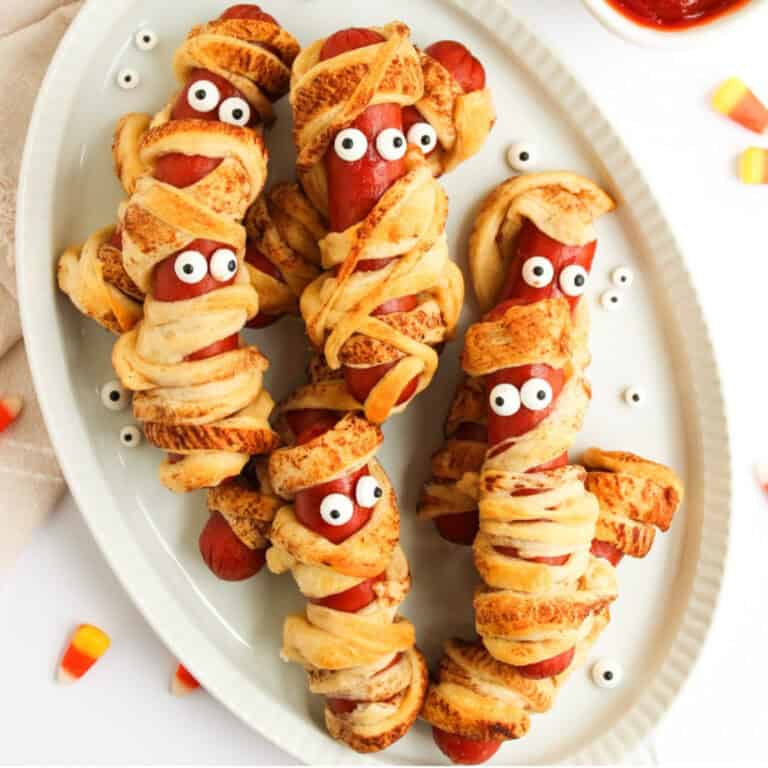 Hot dogs wrapped in crescent roll dough to look like mummies, baked golden brown with candy eyes, arranged on a white platter for Halloween.